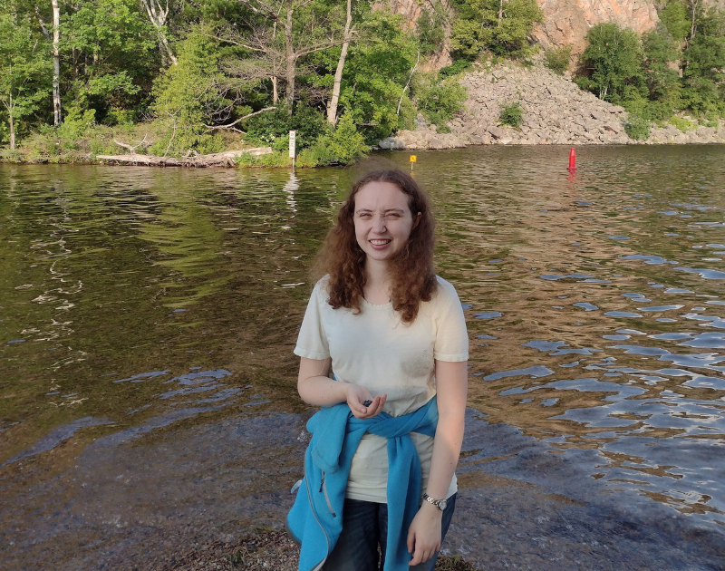 Eleanor stands in front of a lake and cliffs, wearing a light green t-shirt with a blue fleece tied around the waist.