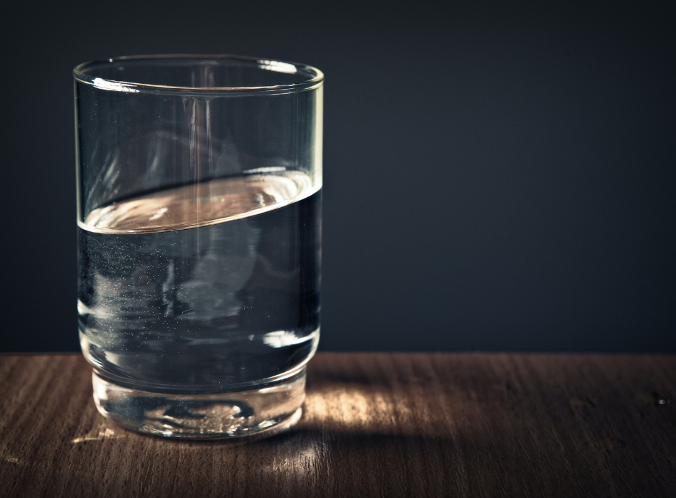 A half full glass of water sits on a counter in front of a dark grey wall