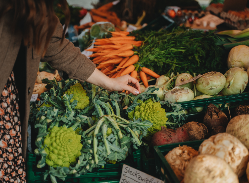 A woman wearing a flower-print dress and brown coat checks the quality of a romanesco that's sitting among other bins of produce