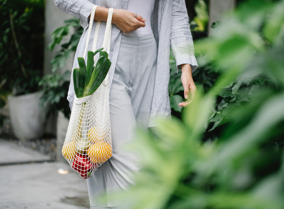 A woman dressed in pale grey carries a cotton mesh bag full of fruit and vegetables