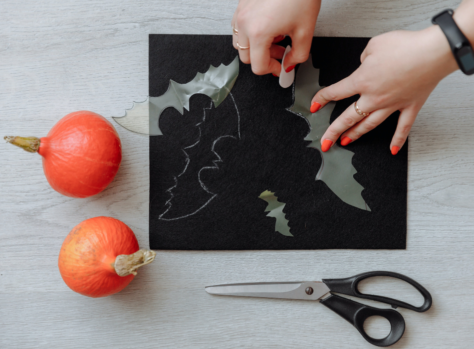 A woman's hands wearing orange nail polish pull cutout bats out of construction paper. A pair of scissors is below the paper, and two small orange pumpkins are to the left.