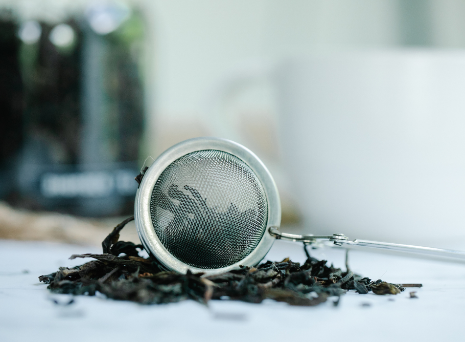 A mesh tea strainer rests on a pile of loose tea leaves that are spilled across a white counter.