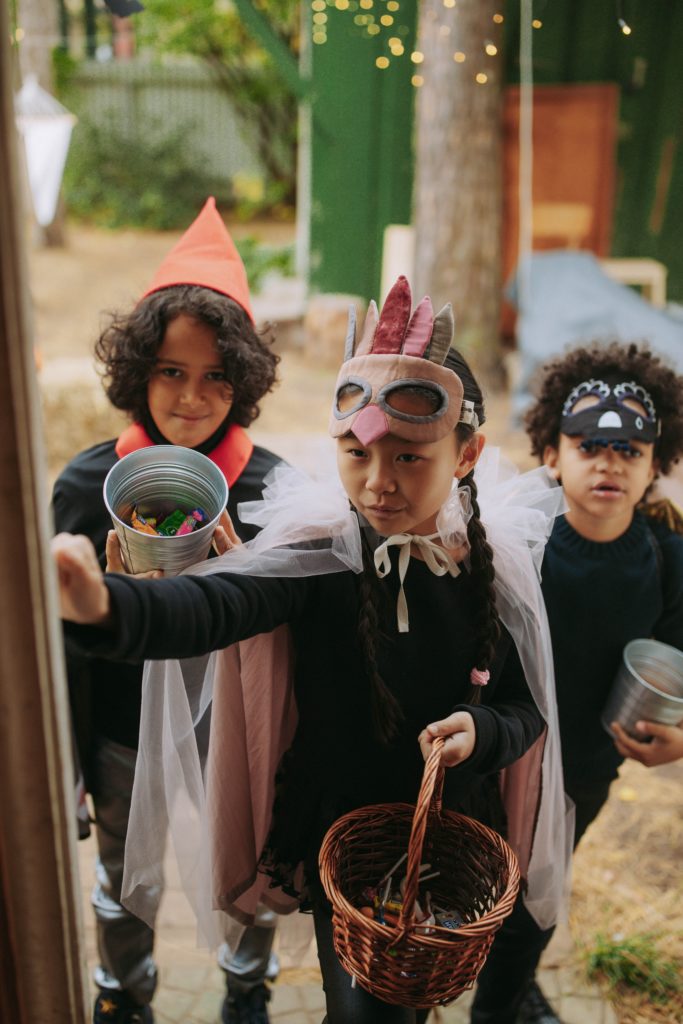 Three children in costume knock at a door, carrying baskets and buckets of candy.