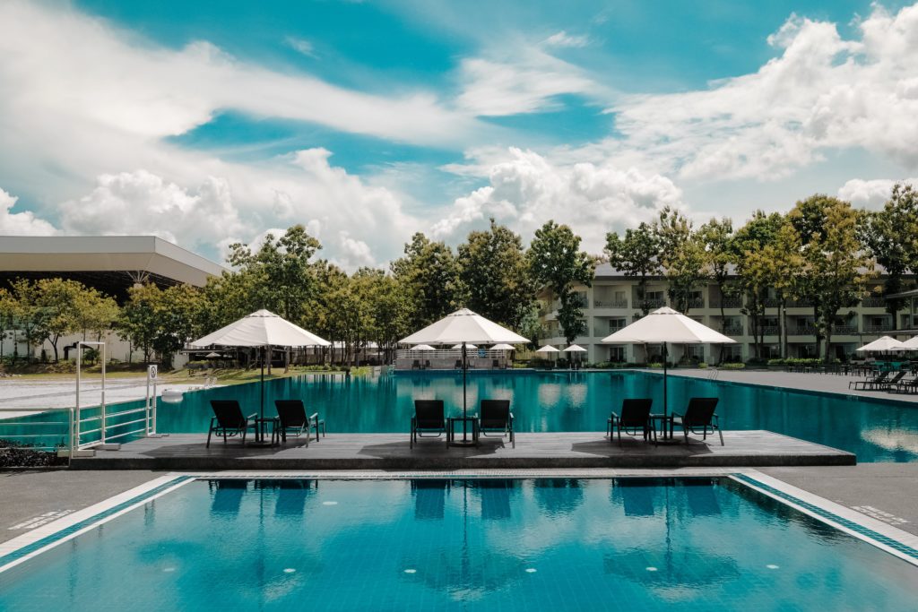 On a sunny day, three sets of chairs under beach umbrellas overlook the turquoise waters of a pool, as well as trees and a white low-rise building on the far side of the water.