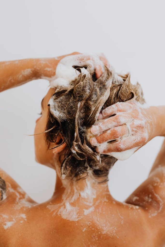 A woman is seen from the backbones up, lathering shampoo in her brown hair.