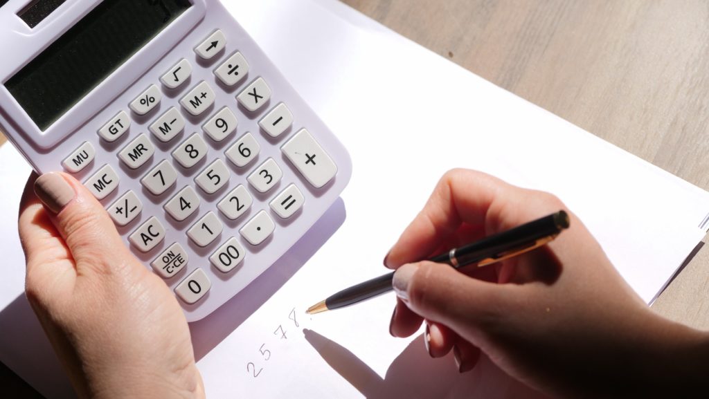 A woman's left hand holds a white calculator, while her other hand writes calculations on a piece of paper