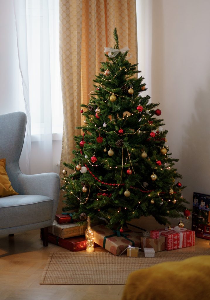 A Christmas tree decorated with colourful balls and garlands is set up in a modern living room. At its base are presents wrapped in red and gold wrapping paper.