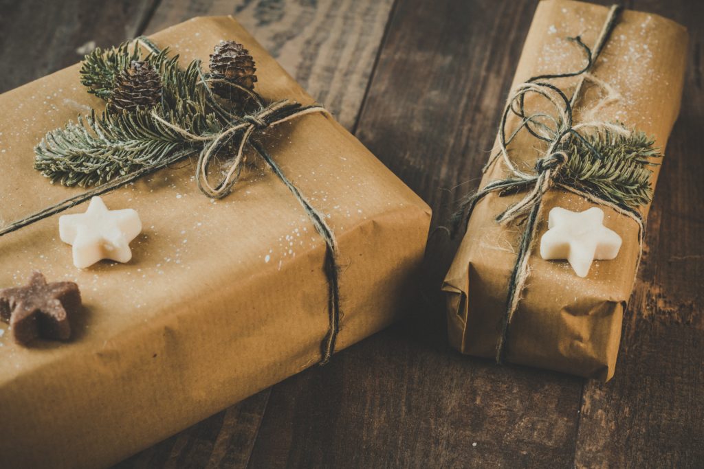 Two presents wrapped in brown paper and string and decorated with pine twigs and carved stars sit on a wooden surface.