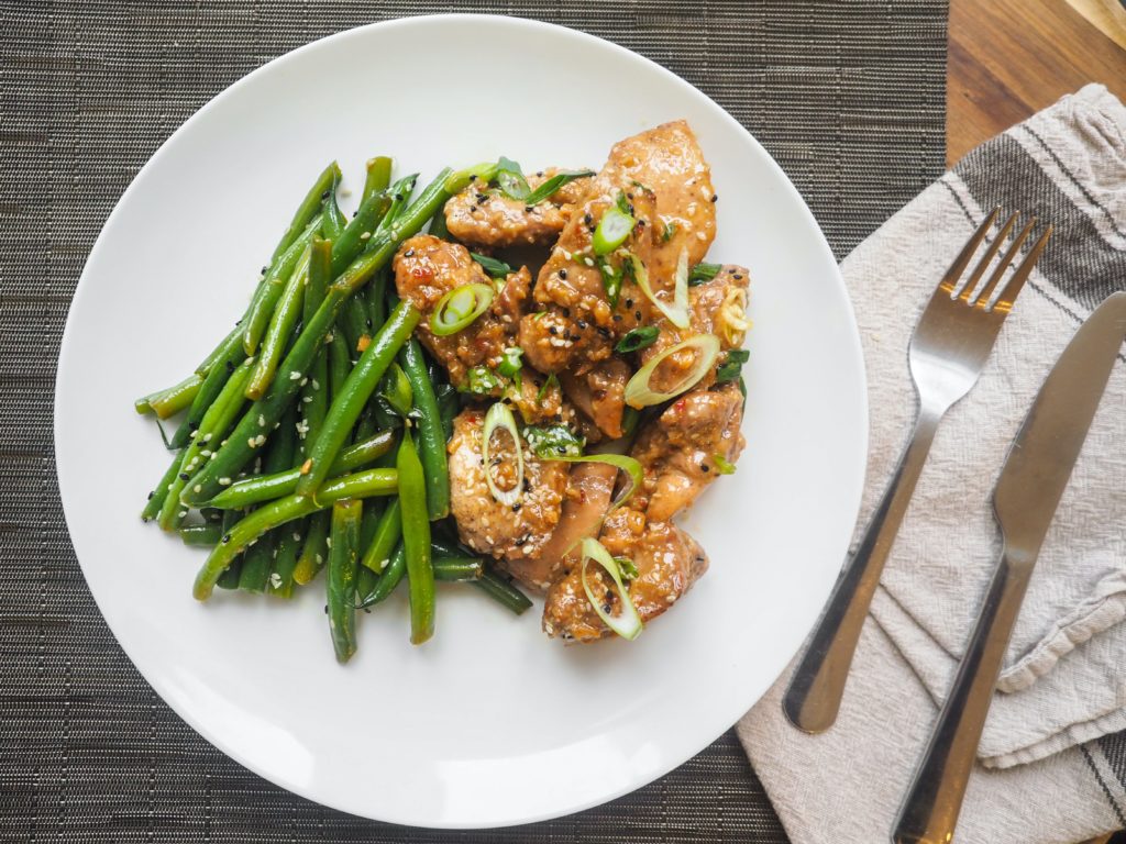 A white plate contains a meal of green beans and pieces of chicken in a sauce. To the right of the plate are a cloth napkin, fork and butter knife