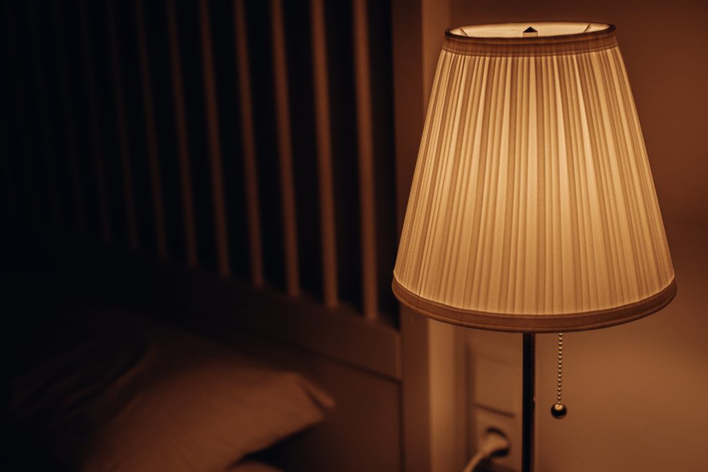 A lit bedside lamp with a light-brown shade stands out in front of a wooden bed frame, which fades into darkness.