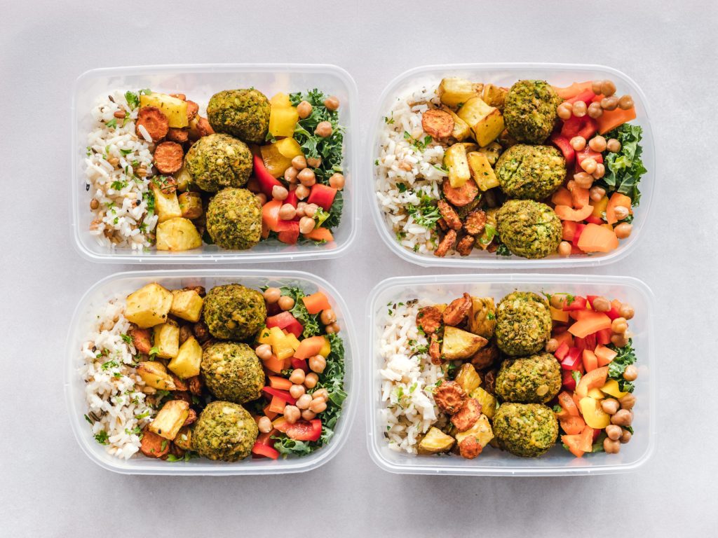 Four clear reusable containers arranged in a grid contain rice, roasted root vegetables, falafels and a colourful salad