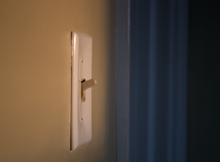 A white light switch is switched on on a light brown wall. A medium-blue curtain hangs in the background.