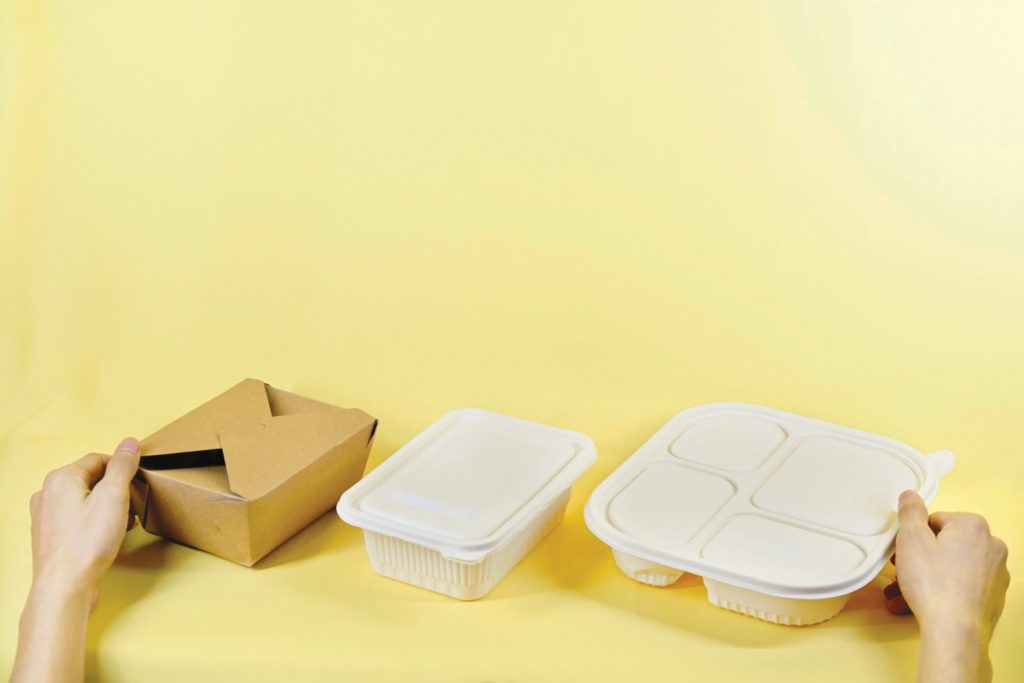 Three takeout containers sit in a row, one made of paper and the other two of plastic. There are hands holding two of the containers, and they're on a yellow background.