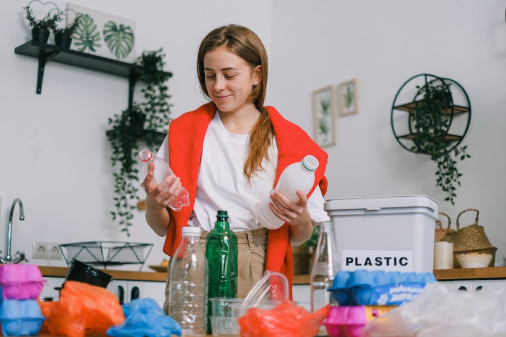 A woman with brown hair wearing a white t-shirt with a bright red scarf looks down at two plastic bottles that she holds in her hands. On the surface in front of her are various kinds of plastic packaging and a bin that says Plastic.