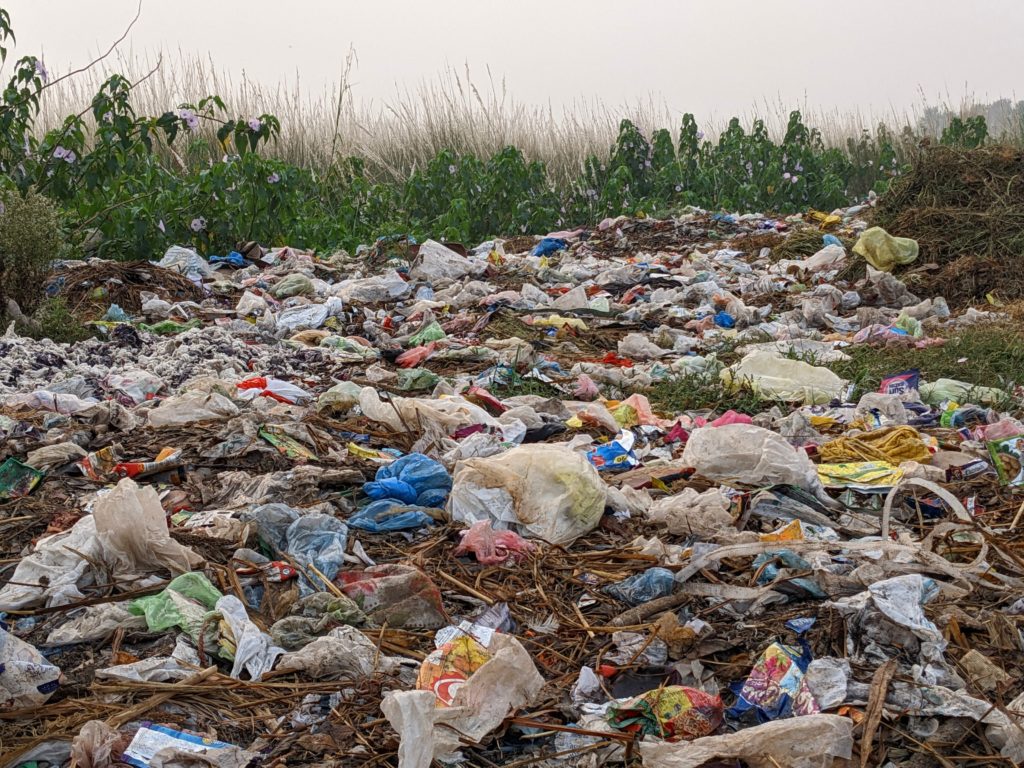 Plastic bags and other pieces of packaging litter an open area with bushes and grasses in the background.