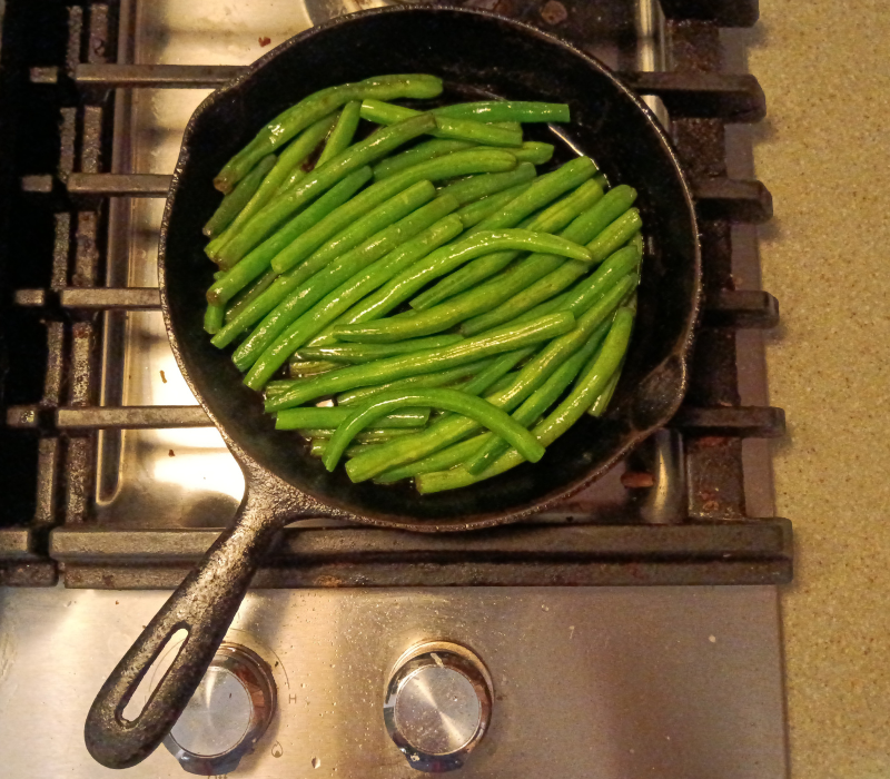 A small cast iron frying pan full of fried green beans sits on the grill of a stove.