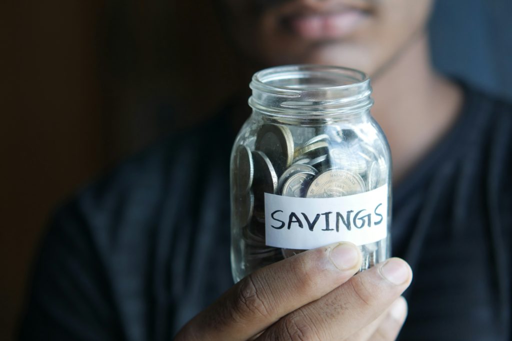 A man holds out a jar full of coins that that says "Savings" on it.