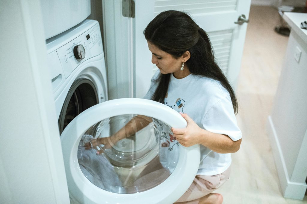 A woman with dark brown hair wearing a white t-shirt crouches loading clothing into a white, front-loading washing machine.