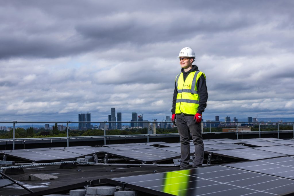 A young man wearing a white hard had and a yellow reflective vest stands on top of a roof with solar panels on it.