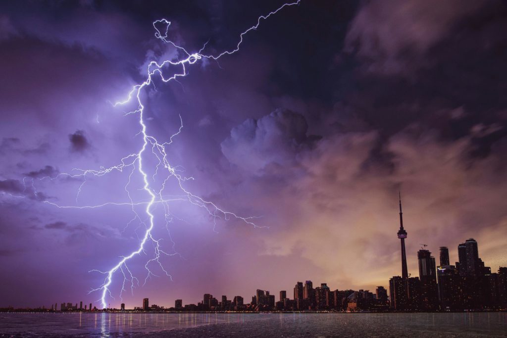 A large lightning bolt strikes buildings in the distance, against a sky purple with storm clouds. In the foreground is a lake and city skyline.