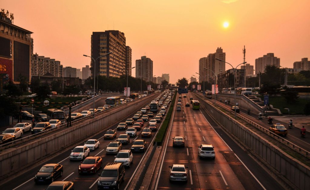 An urban highway with gridlock on one side and lighter traffic going the other direction. There are tall buildings in the background and the sky is hazy.