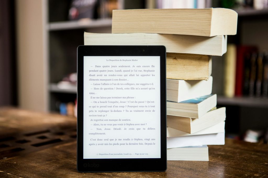 An e-reader with text on it stands in front of a pile of books.