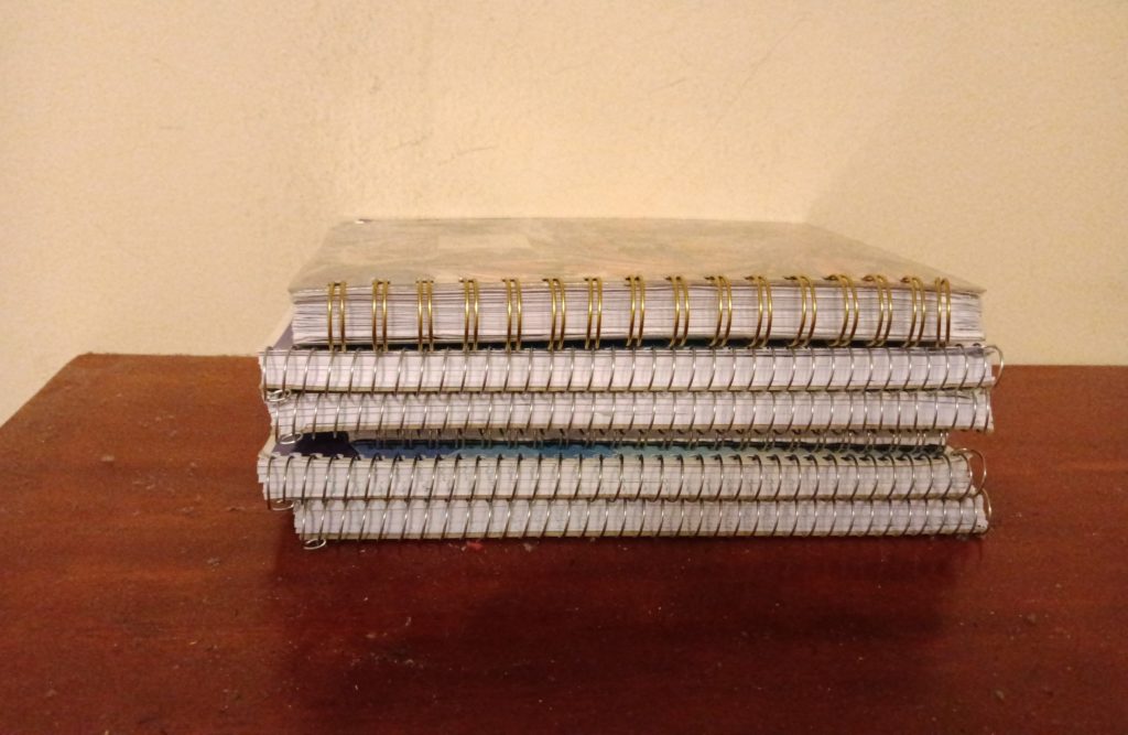 A stack of coil notebooks sits on top of a wooden desk.