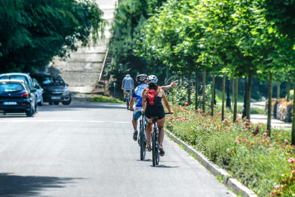 A woman and man bike single file down a quiet street with a row of trees off to their right.