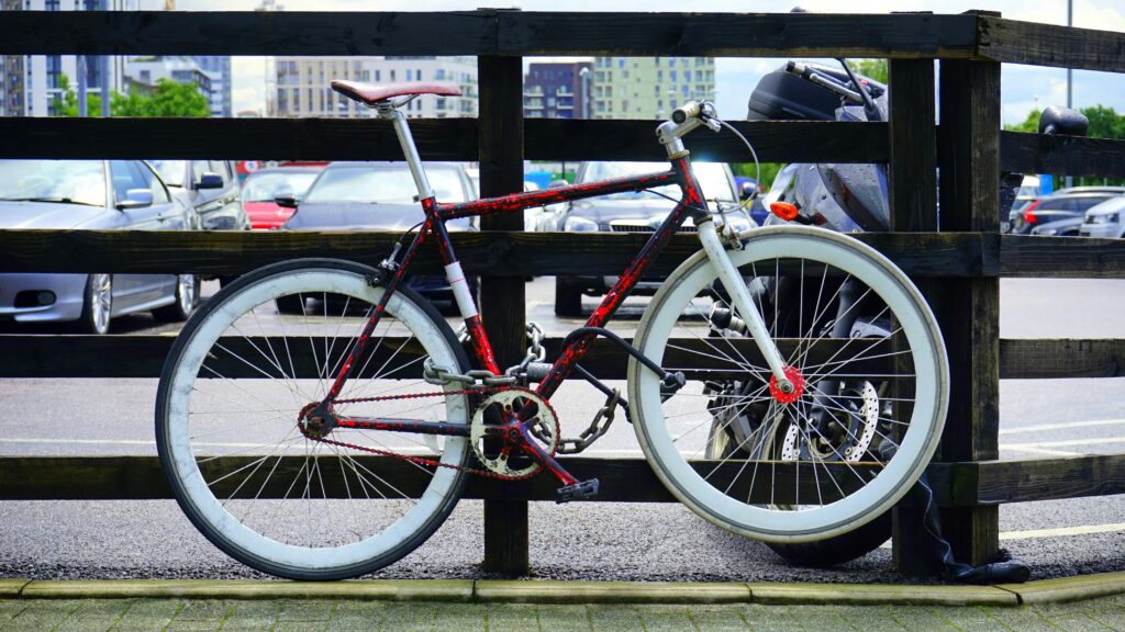 A red bicycle with white rims is chained up to a wooden fence outside a parking lot.