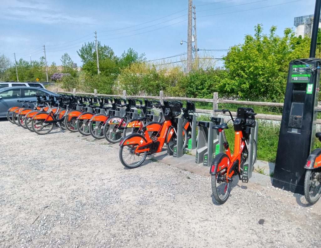 A station of docked orange bicycles sits in a gravel parking lot.