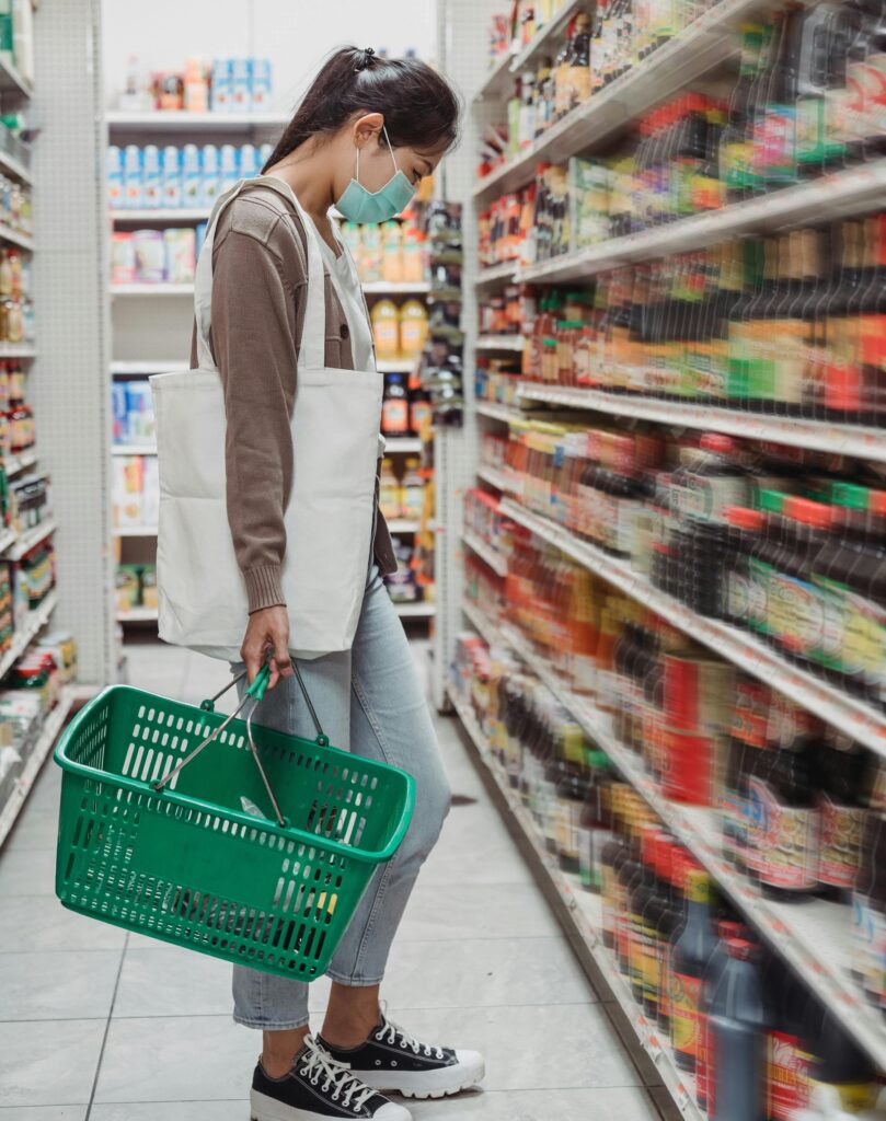 A young woman wearing a mask, a brown long-sleeved shirt and jeans stands in the sauce aisle of a grocery store carrying a white reusable bag and a green shopping basket.