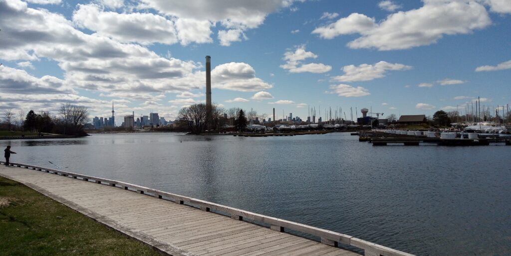 A boardwalk follows a calm body of water with boats, industry and skyscrapers in the background.