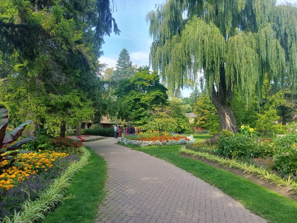 A path cuts through the centre of a garden with colourful flower beds, green grass and weeping willows on either side.