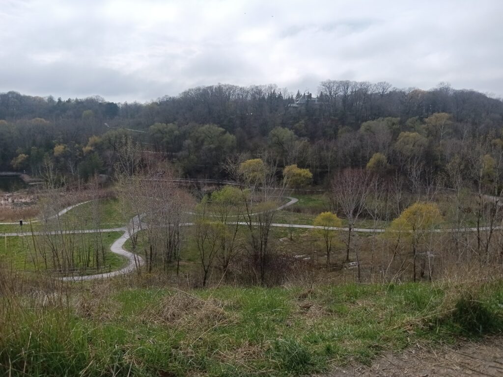 Walking trails crisscross green space at the foot of a cliff with trees in the background.