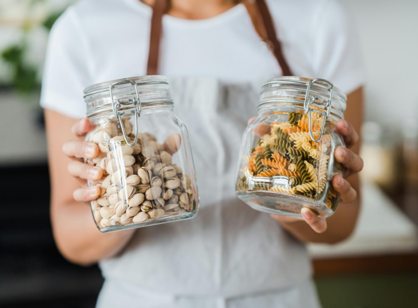 A woman wearing a white t-shirt holds two glass jars containing pistachios and dried pasta.