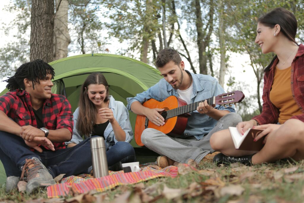 A group of four young adults sit around a picnic blanket, in front of a tent.