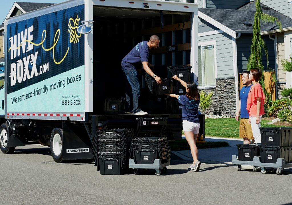 A man in the back of a truck hands a black plastic bin down to a woman. There are stacks of more bins on the street.