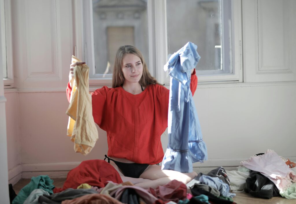 A woman wearing a red shirt holds up two items of clothing, surrounded by piles of clothing on the floor.