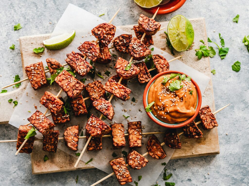 Skewers with chunks of tempeh sit on a chopping board along with an orange sauce.