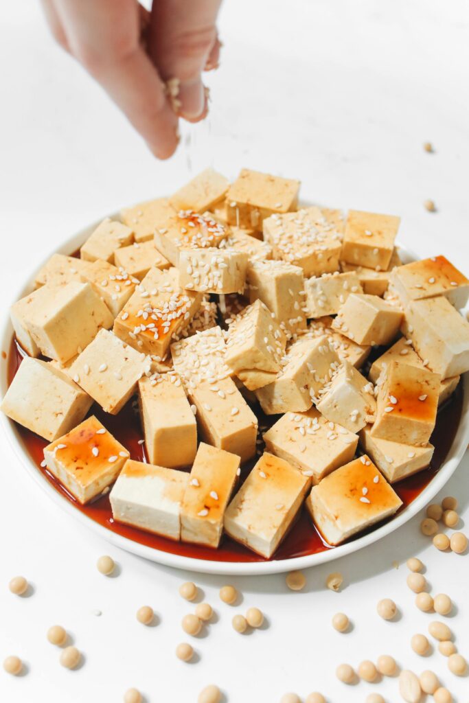 A person sprinkles sesame seeds onto a bowl of tofu with sauce.