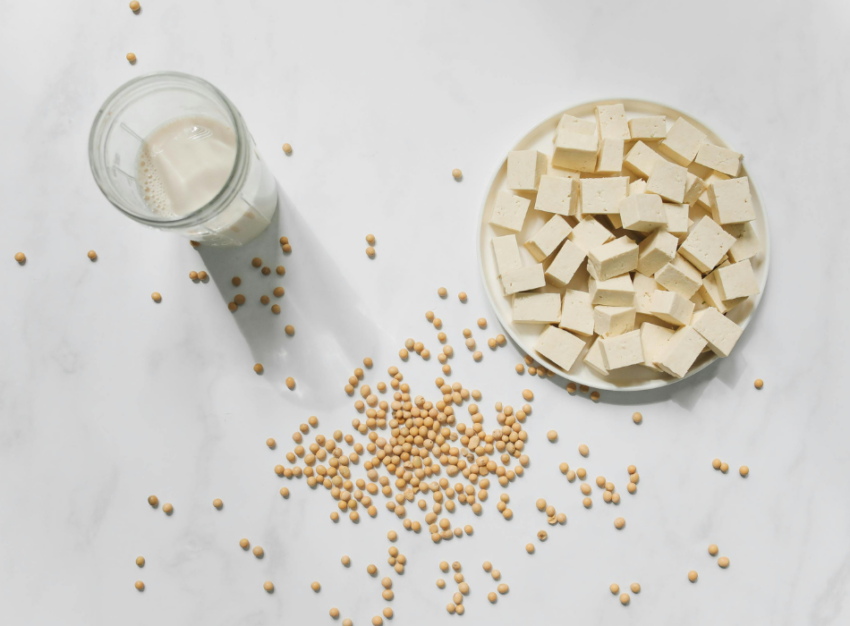 A bowl of tofu cubes sits next to scattered soybeans and a glass of soymilk.