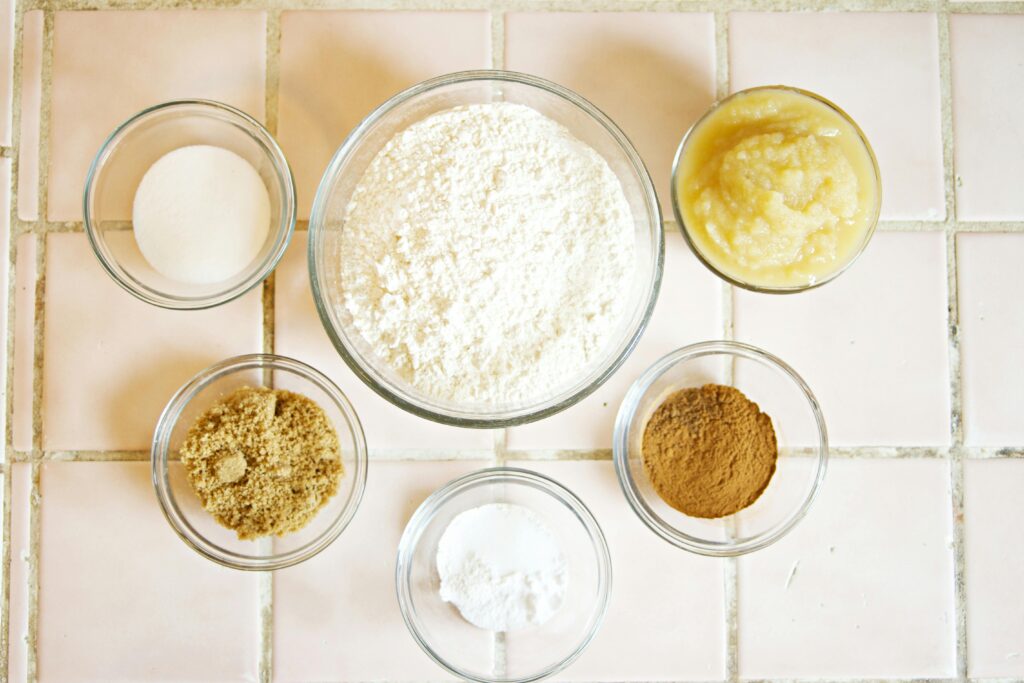 A set of small baking bowls contain flour, spices and apple sauce.