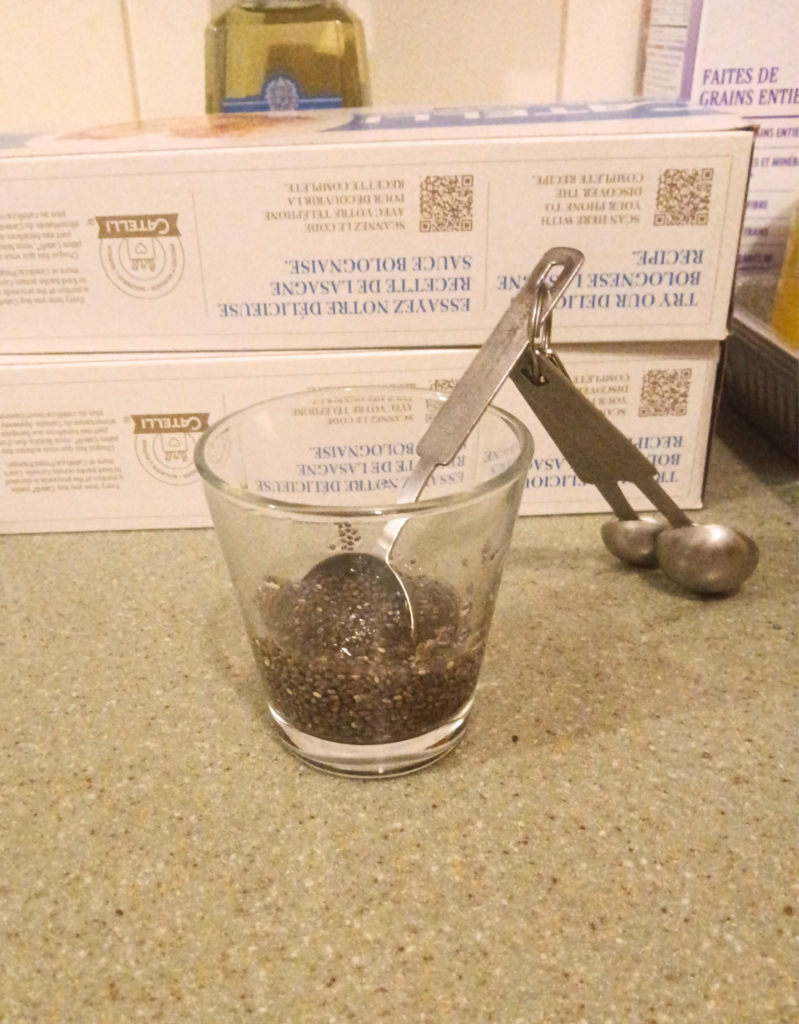 A clear glass contains chia seeds mixed with water with a set of measuring spoons sticking out.