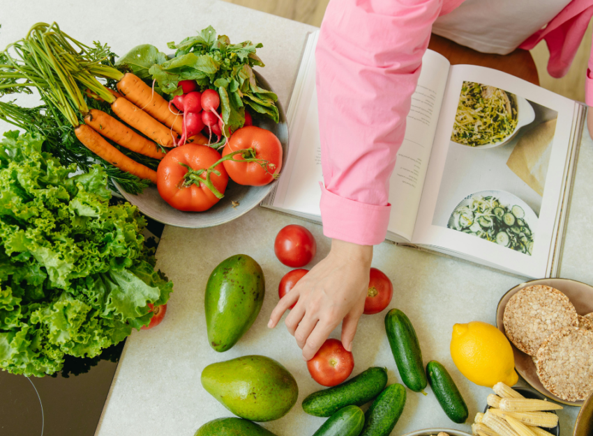 An arm reaches across an open cookbook to a group of colourful vegetables sitting on a counter.