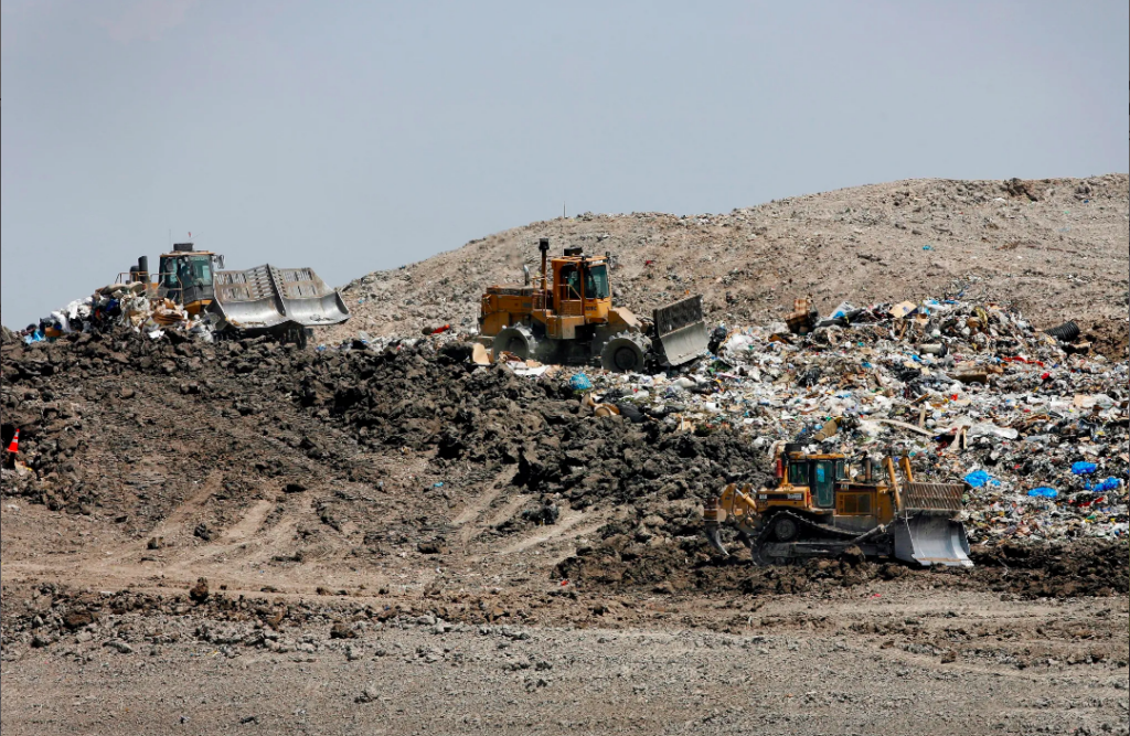 Three bulldozers drive over a pile of trash at a landfill.