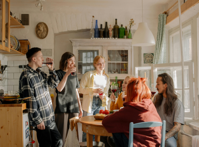 Six roommates gather around a round dining table, drinking red wine.