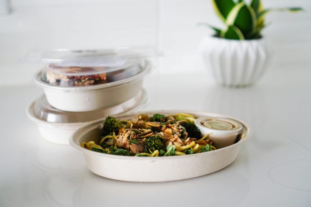 A round takeout container with a vegetable stir-fry, with other containers and a potted plant in the background.