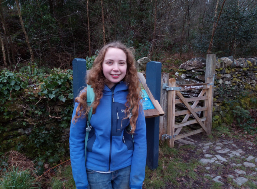 A girl with curly hair wearing a backpack poses in front of the start of a walking trail.